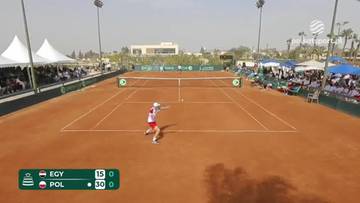 Gracz w stroju tenisowym odbija piłkę na korcie ziemnym. W tle widownia, namioty i budynki.