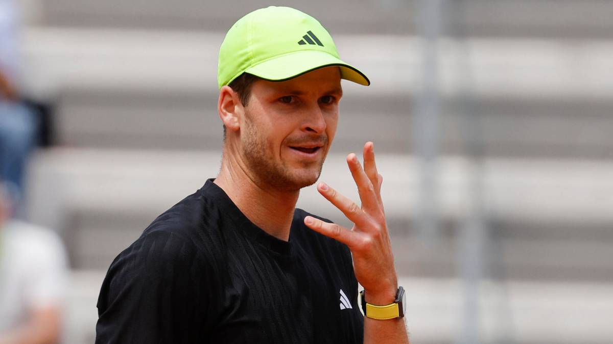 Hubert Hurkacz w żółtej czapce i czarnej koszulce, z podniesionymi trzema palcami prawej dłoni.