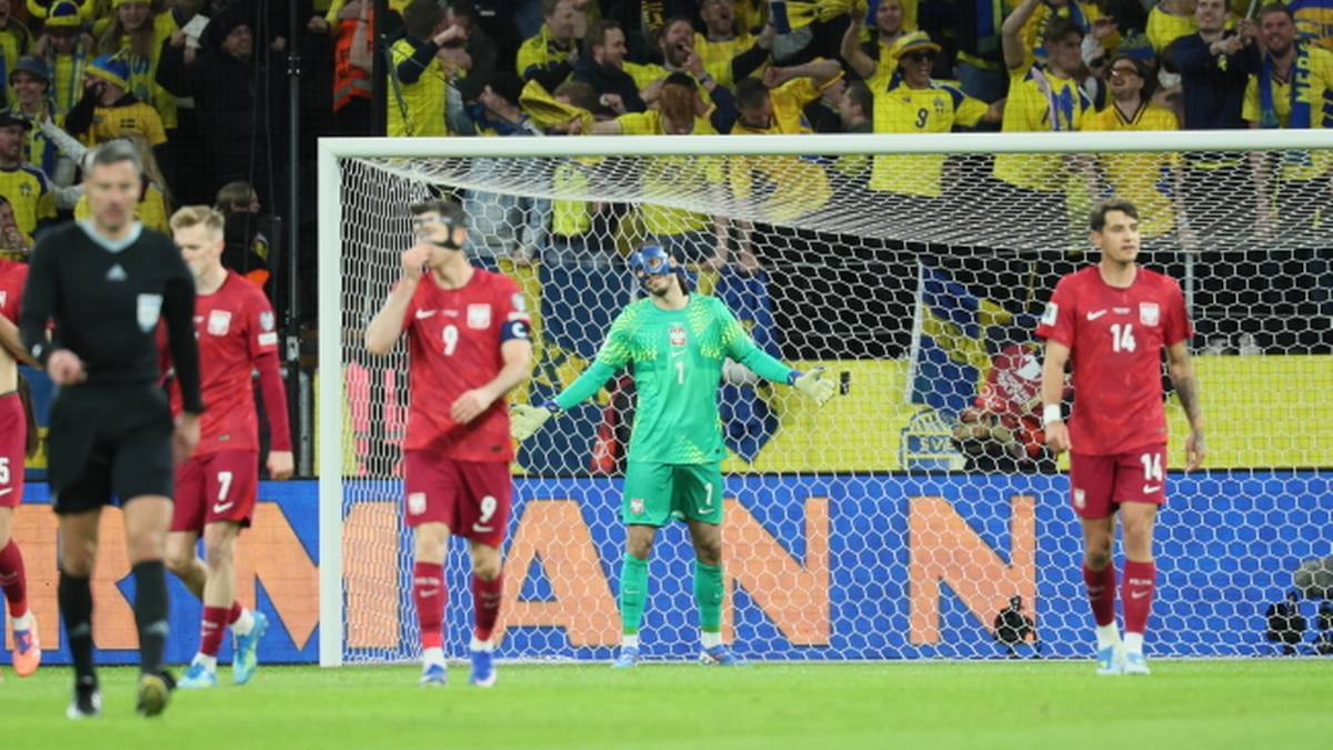 Bramkarz w zielonym stroju stoi na boisku piłkarskim, otoczony przez zawodnik&oacute;w w czerwonych koszulkach. W tle widoczna jest bramka i trybuny z kibicami.