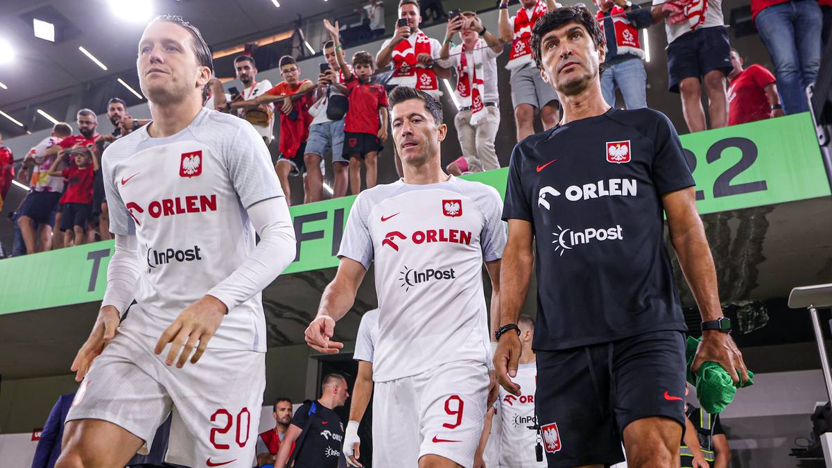 Trzech piłkarzy reprezentacji Polski w strojach meczowych na tle trybun z kibicami.