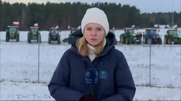 Młoda kobieta w białej czapce i ciemnej kurtce z mikrofonem przed aparatem, na tle szeregu traktor&oacute;w i flag.