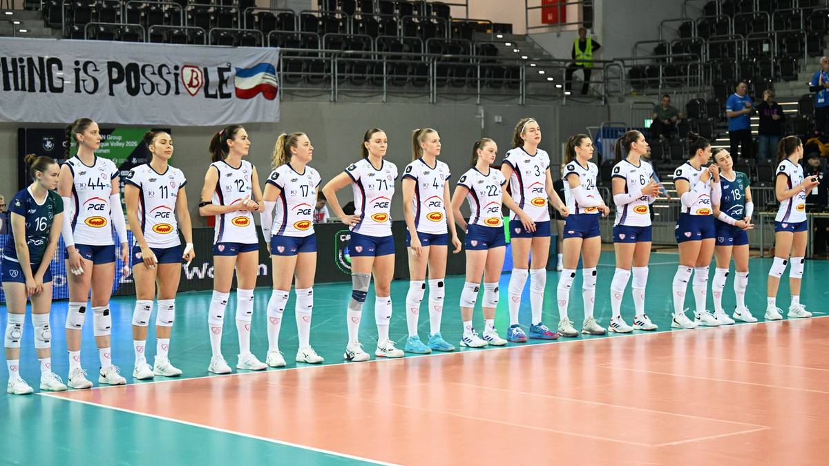 Drużyna siatkarek w białych strojach sportowych stoi w rzędzie na boisku.