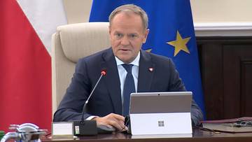 Premier Donald Tusk siedzi przy stole, przed nim laptop, za nim flagi Polski i Unii Europejskiej.