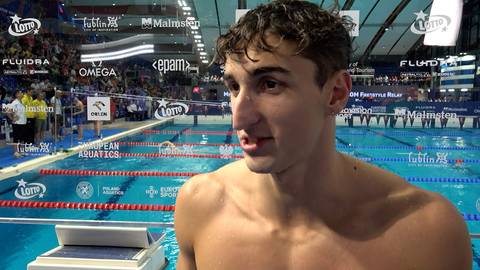 Portret młodego mężczyzny, pływaka, w zbliżeniu. W tle widać basen pływacki z torami i trybunami.