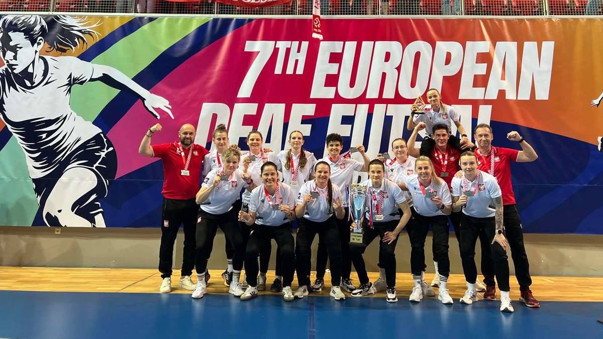 Drużyna polskich piłkarek pozuje do zdjęcia z flagą Polski w tle, świętując sukces na Mistrzostwach Europy w Futbolu Os&oacute;b Niesłyszących.
