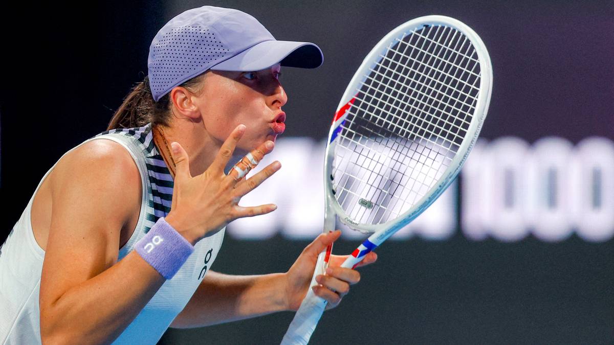 Iga Świątek w czapce i bluzce tenisowej, dmucha na dłoń trzymając rakietę tenisową.