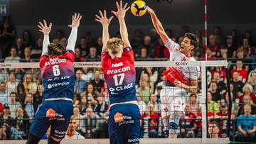 Siatkarze w akcji przy siatce, jeden z nich atakuje piłkę, pozostali pr&oacute;bują ją zablokować.