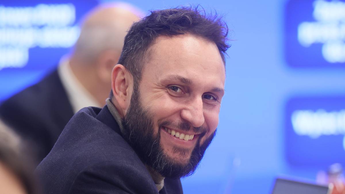 Uśmiechnięty mężczyzna z brodą siedzi przy stole konferencyjnym, w tle widoczne inne osoby i laptopy.