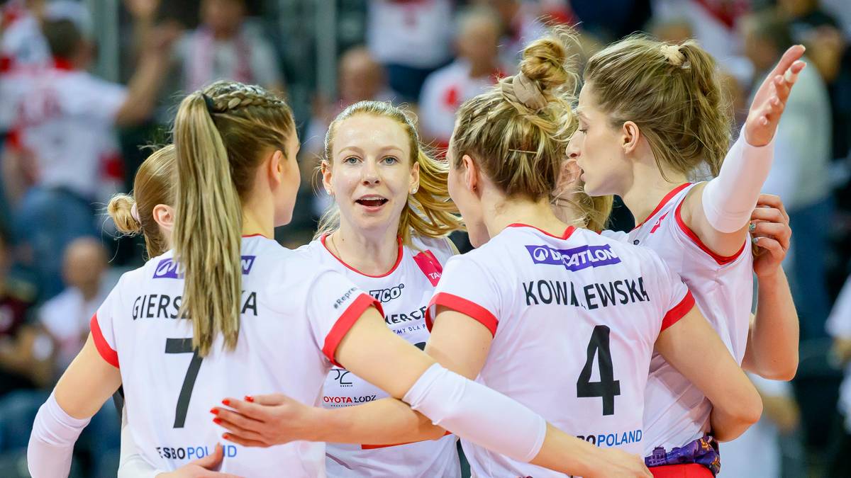 Grupa siatkarek w białych strojach z czerwonymi elementami stoi w kółku, przytulając się. Widoczne numery na koszulkach: 7 i 4. Uśmiechy na twarzach.