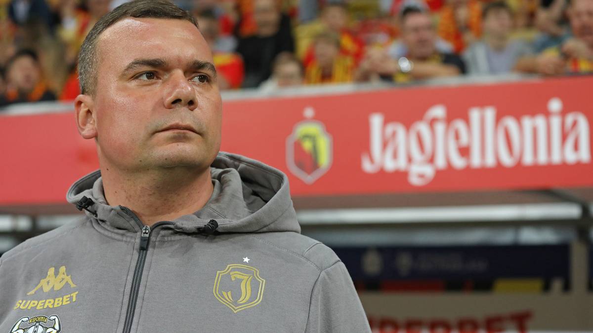 Portret trenera piłkarskiego Jagiellonii Białystok w szarej bluzie z kapturem. W tle widać trybuny z kibicami i baner z logo klubu.