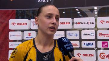 Młoda kobieta w ż&oacute;łtej koszulce sportowej z mikrofonem przed twarzą, na tle ścian z logotypami sponsor&oacute;w.