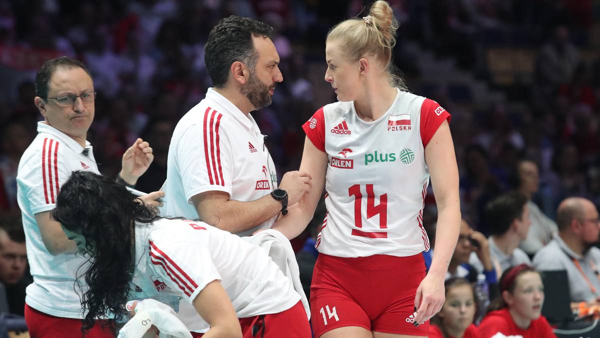 Trener rozmawia z zawodniczką podczas meczu siatk&oacute;wki, w tle widoczni są inni członkowie sztabu i kibice.