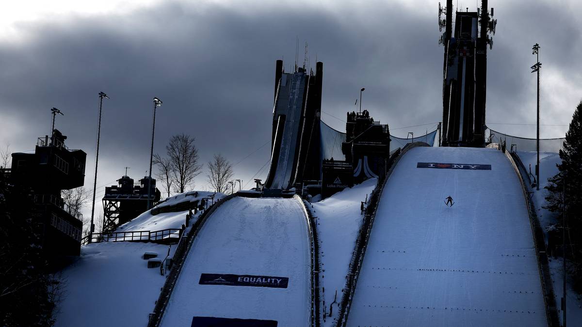 Szeroki kadr przedstawiający zimowy krajobraz ze skocznią narciarską. Na pierwszym planie śnieżne zeskoki, a w tle widoczne wysokie wieże startowe i konstrukcje budynków.