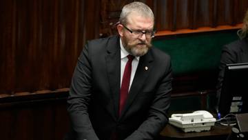 Grzegorz Braun w garniturze i okularach stoi przy mównicy.