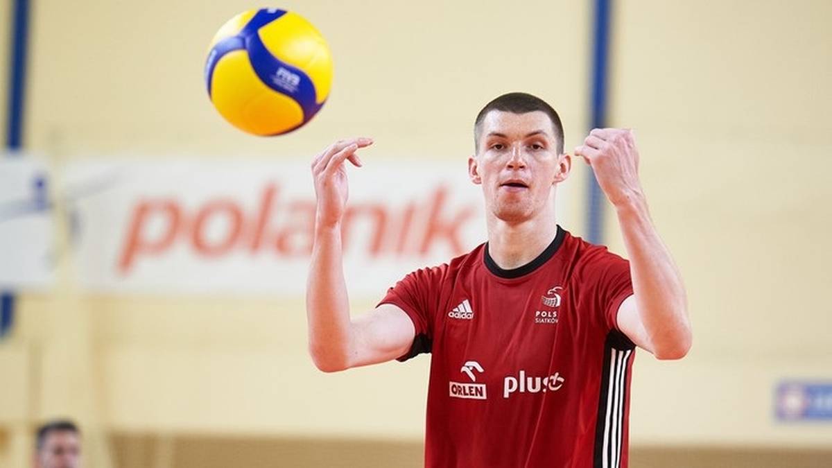 Młody siatkarz w czerwonej koszulce ćwiczy zagrywkę. Piłka do siatk&oacute;wki unosi się nad jego głową.