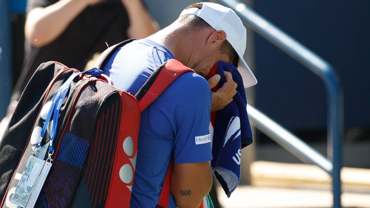 Tenisista z białą czapką i niebieską koszulką opiera się o ręcznik, którym wyciera twarz. Na plecach ma plecak z czerwonymi elementami.