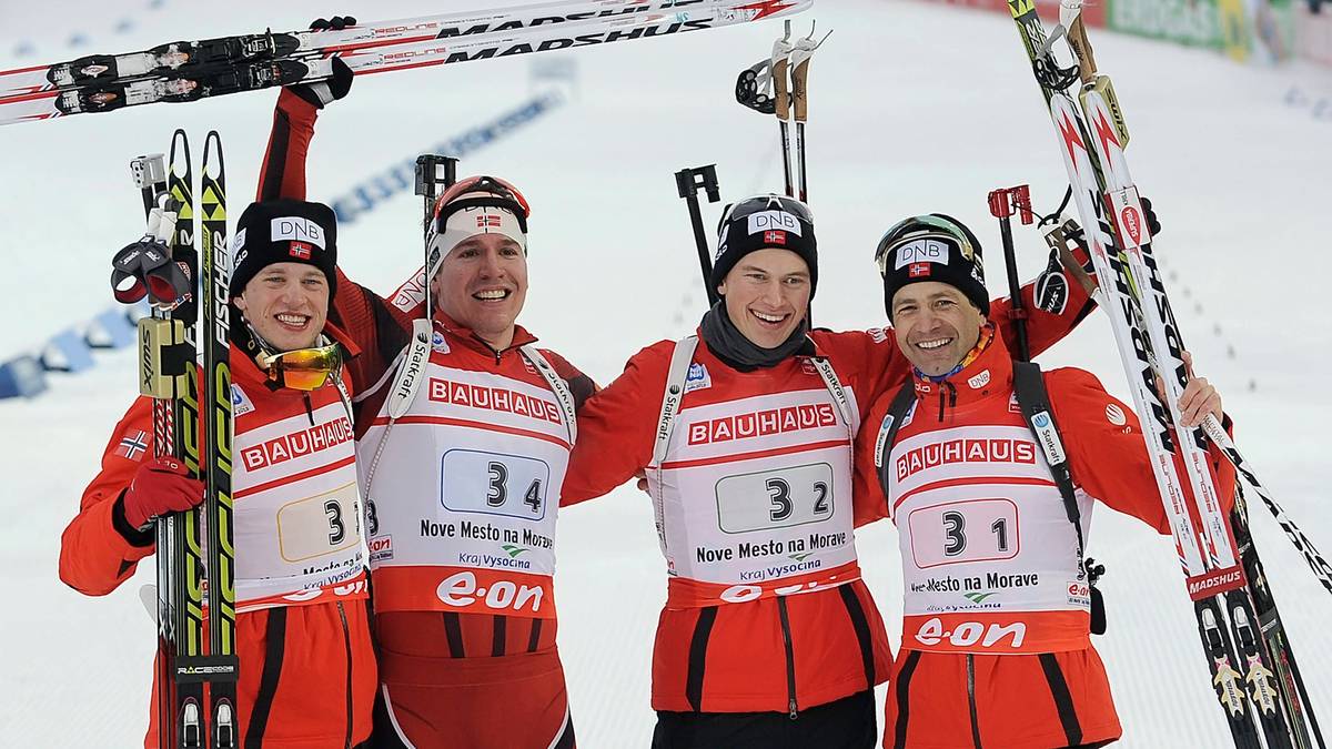 Czterech biathlonistów w pomarańczowo-białych strojach sportowych pozuje do zdjęcia z nartami. Wszyscy uśmiechają się i patrzą w stronę obiektywu.