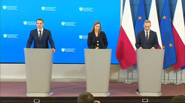 Trzy osoby, dw&oacute;ch mężczyzn i jedna kobieta, stoją za m&oacute;wnicami na tle niebieskiej ścianki z logo. Konferencja prasowa.