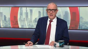 Mężczyzna w okularach, koszuli i krawacie siedzi za stołem z dokumentami i kubkiem, patrząc na widza. Tło stanowi ekran z widokiem na miasto.