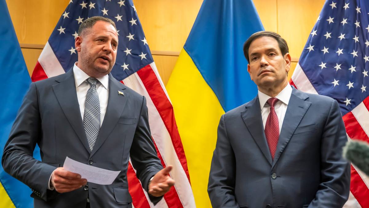 Dwóch mężczyzn w garniturach stoi przed flagami USA i Ukrainy, symbolizując spotkanie delegacji obu krajów.