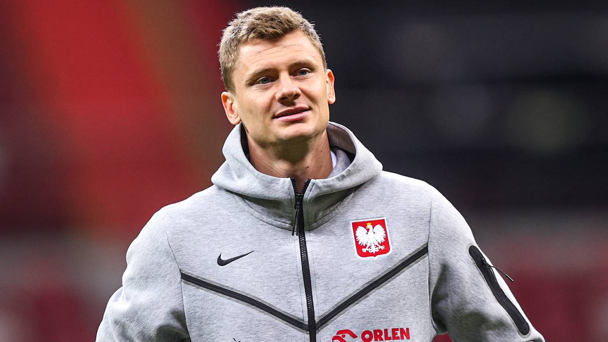 Portret piłkarza w szarej bluzie z kapturem z herbem Polski na piersi.