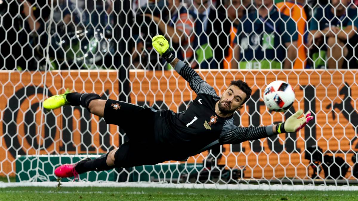 Bramkarz w czarnym stroju z numerem 1 na plecach broni strzał, rzucając się w kierunku piłki, która leci w stronę bramki.