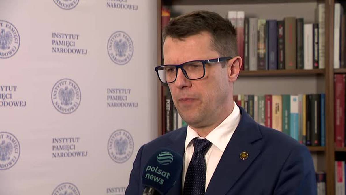 Mężczyzna w okularach i garniturze, stojący przed ścianą z logo Instytutu Pamięci Narodowej. Trzyma mikrofon Polsat News.
