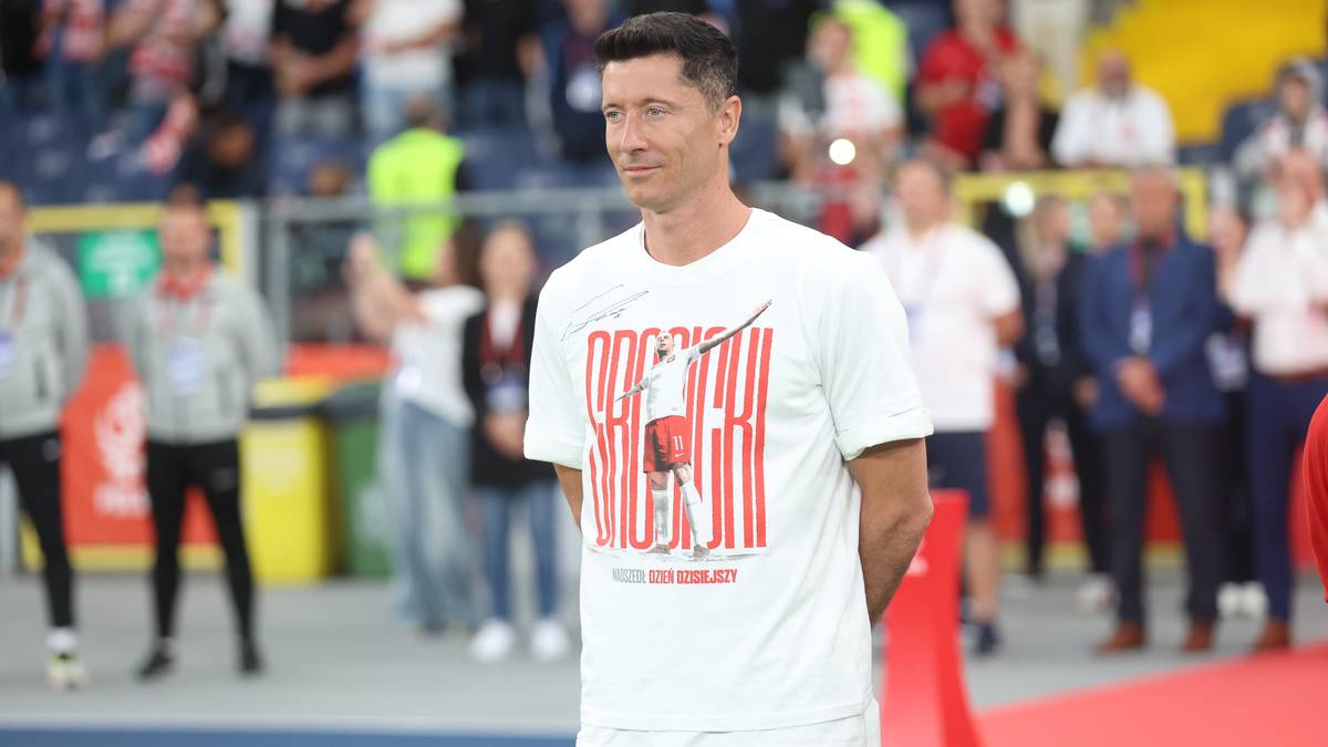 Robert Lewandowski w białej koszulce z czerwonymi napisami i grafiką, stoi na boisku piłkarskim.