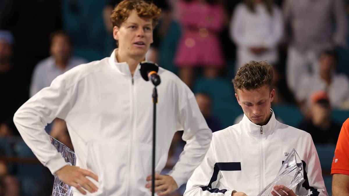 Dw&oacute;ch tenisist&oacute;w w białych strojach sportowych na podium podczas ceremonii wręczenia nagr&oacute;d. Jeden z nich, z pucharem w ręku, patrzy w d&oacute;ł, drugi stoi obok niego, patrząc w stronę mikrofonu.