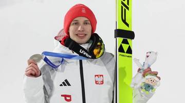 Młody skoczek narciarski w czerwonej czapce i białej kurtce z orzełkiem na piersi, trzyma srebrny medal i narty z logo FIS.