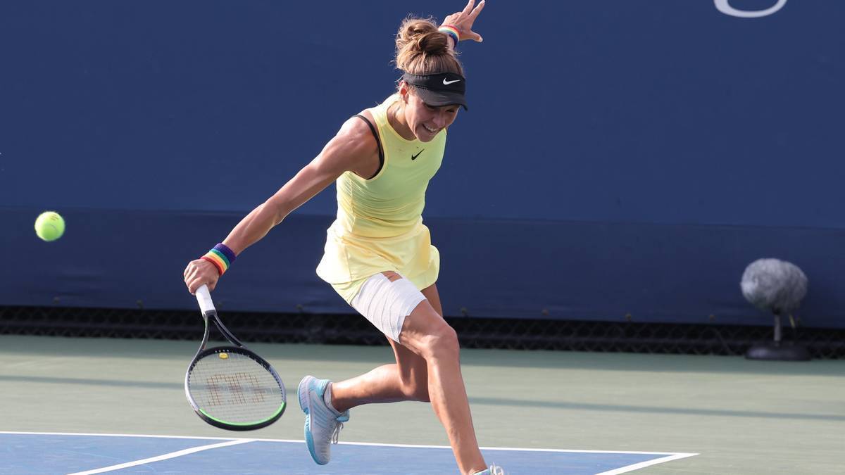 Tenisistka w żółtej sukience i białych spodenkach wykonuje forhend podczas meczu tenisowego na niebieskim korcie.