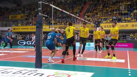 Gracz w niebieskim stroju z numerem 10 skacze do ataku podczas meczu siatk&oacute;wki. Gracze w ż&oacute;łtych strojach po drugiej stronie siatki obserwują akcję. Widownia na trybunach w tle.