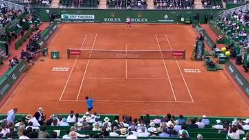 Kort tenisowy z mączki ceglanej podczas meczu, z zawodnikiem w niebieskiej koszulce odbijającym piłkę i widzami na trybunach.