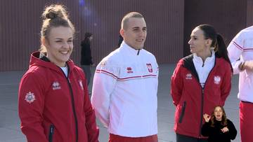 Trzy osoby ubrane w sportowe stroje, dwie kobiety i jeden mężczyzna, stoją obok siebie na zewnątrz. Dwie kobiety mają na sobie czerwone kurtki z białymi elementami, a mężczyzna białą kurtkę z czerwonymi i białymi pasami. W prawym dolnym rogu znajduje się małe okno z tłumaczką języka migowego.