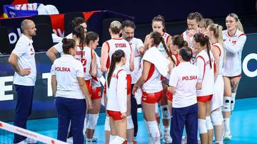 Dyskretnie ustawiona grupa graczy reprezentacji Polski w siatk&oacute;wce, w tym kilka kobiet i jeden mężczyzna stojący z boku, zamyka krąg, aby wysłuchać wskaz&oacute;wek od trenera stojącego na czele grupy.