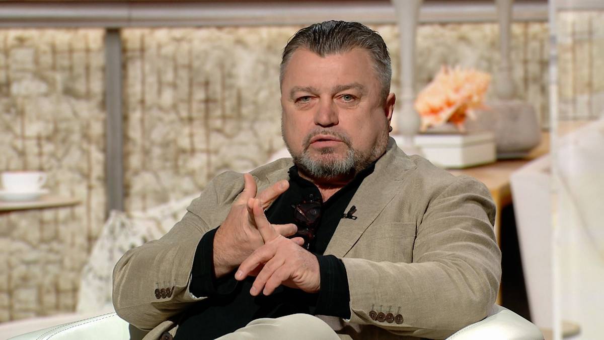 Mężczyzna w garniturze siedzi w fotelu, krzyżując ręce na piersi.