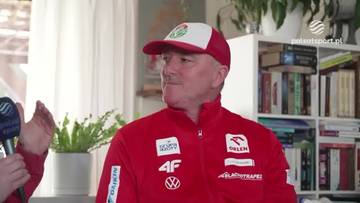 Mężczyzna w czerwonej kurtce sportowej i czapce z daszkiem, ubrany w stroje z logotypami sponsor&oacute;w, udziela wywiadu. W tle widoczne są regały z książkami i zielone rośliny.