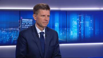 Ryszard Petru w garniturze i krawacie, siedzi na tle niebieskiego ekranu z widokiem na nocne miasto.