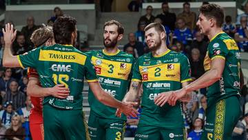 Czterech siatkarzy w zielonych strojach drużyny Aluron CMC Warta Zawiercie rozmawia na boisku, z kibicami na trybunach w tle.