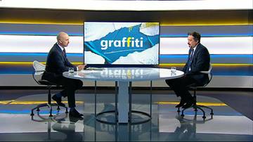 Dw&oacute;ch mężczyzn w garniturach siedzi przy okrągłym stole w studiu telewizyjnym, naprzeciwko siebie. Za nimi znajduje się duży ekran z napisem "graffiti".
