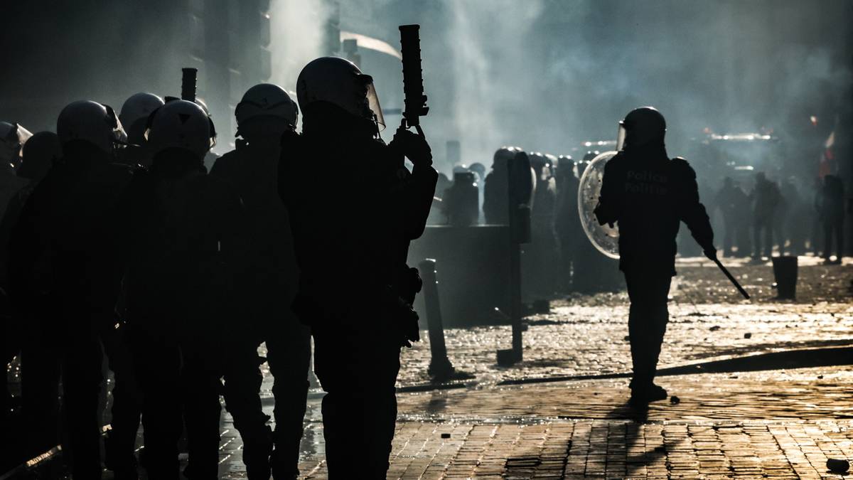 Sylwetki protestujących rolników i policji w dymie podczas zamieszek w Brukseli.