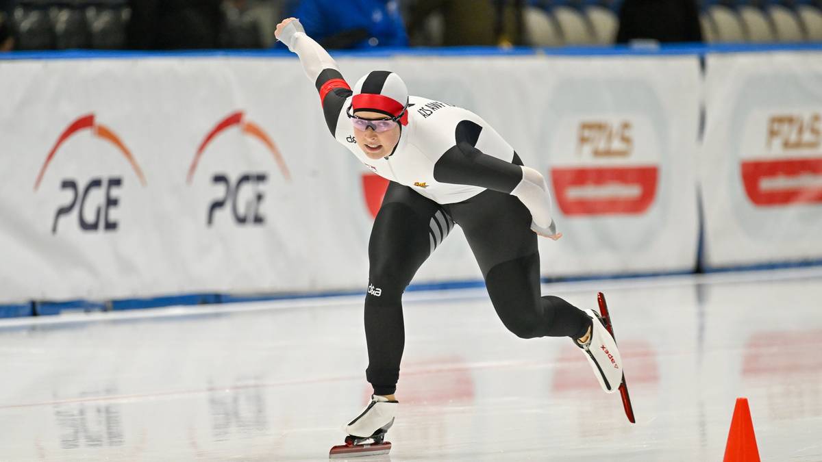 Zawodniczka w białym kombinezonie, kasku i goglach z białymi rękawicami, jedzie na łyżwach po lodowym torze podczas zawodów. Widoczna jest jej sylwetka w ruchu, z uniesioną ręką. W tle znajduje się band a z logo PGE.
