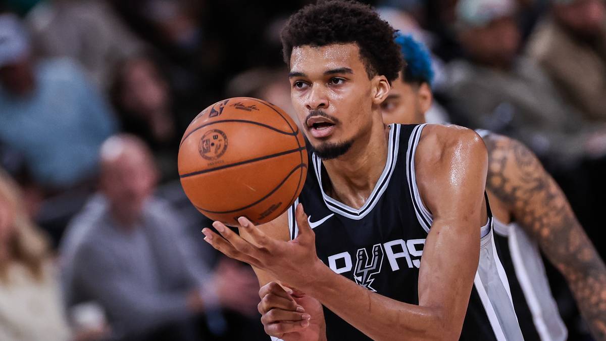 Koszykarz w czarnej koszulce z numerem 10 i białymi paskami, trzymający pomarańczową piłkę do koszykówki. W tle widać rozmazanych kibiców.