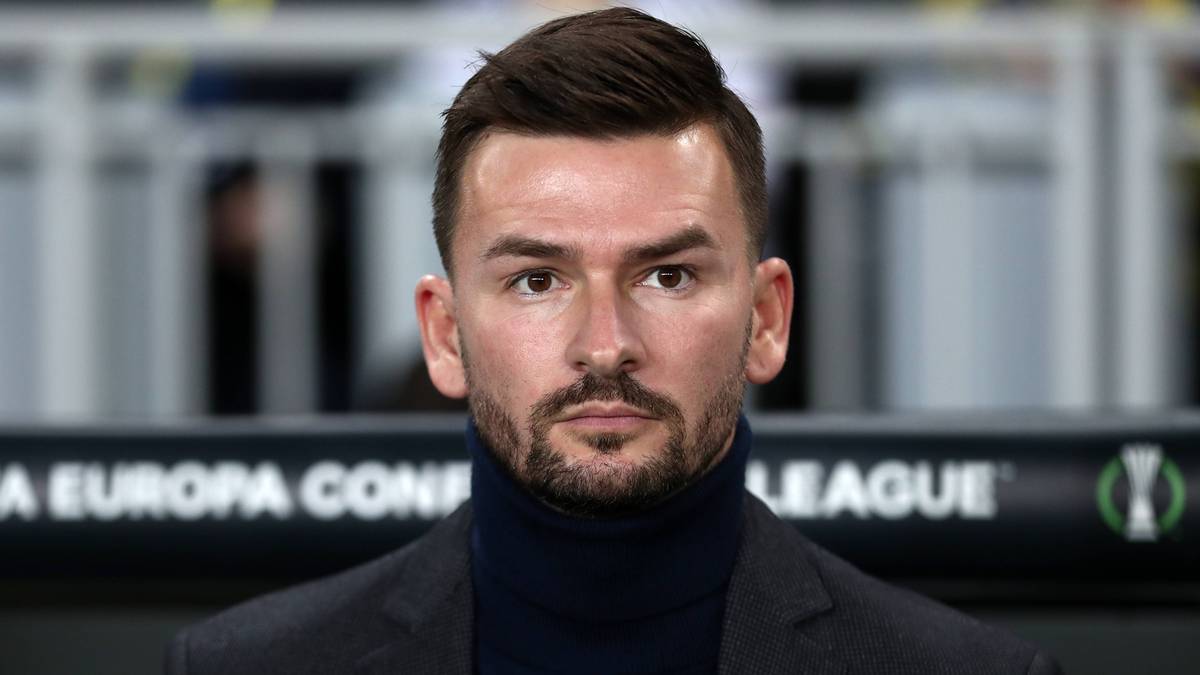 Portret mężczyzny z krótko ostrzyżonymi ciemnymi włosami i zarostem, ubranego w ciemny golf i marynarkę. Tło jest nieostre, widoczne napisy w języku angielskim "EUROPA CONF" oraz "LEAGUE".