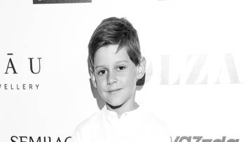 Chłopiec w białej koszuli i jeansach stoi przed ścianką z logotypami.