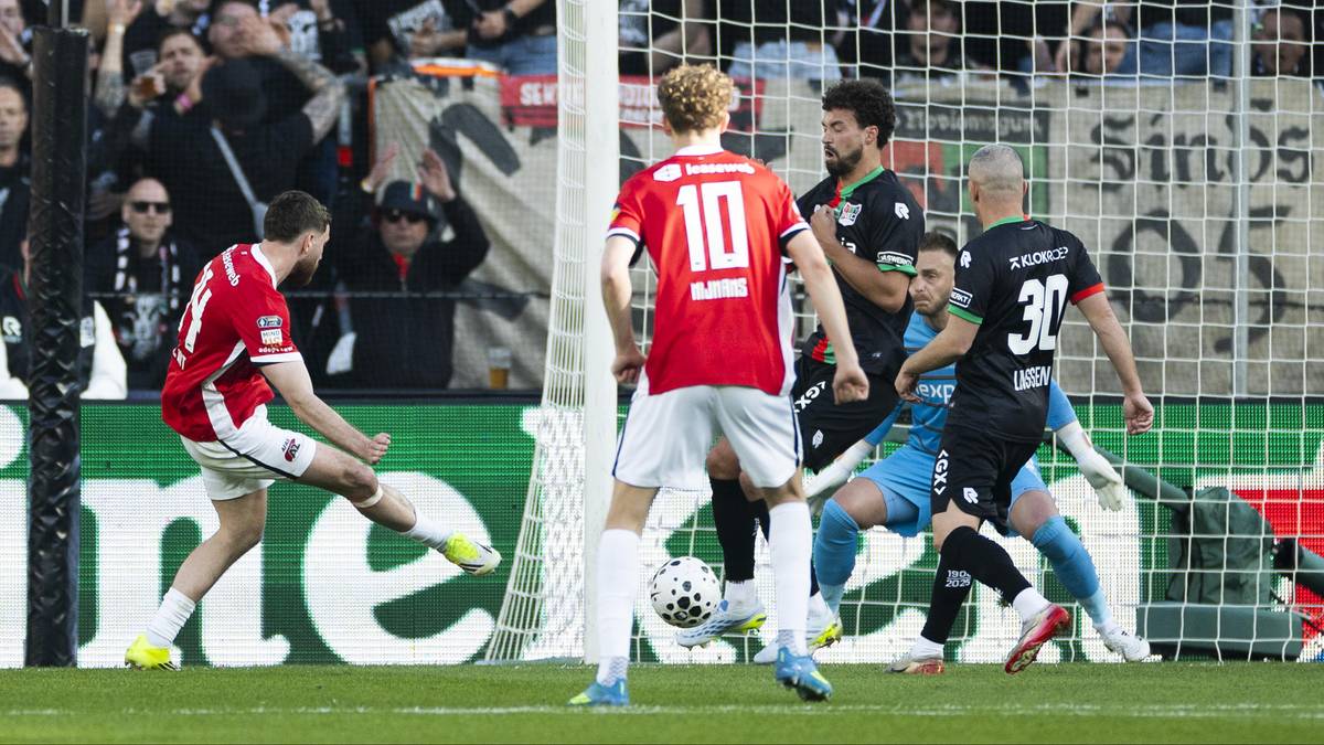 Piłkarze w czerwonych i czarnych strojach walczą o piłkę podczas meczu piłki nożnej. Widzowie na trybunach obserwują grę.