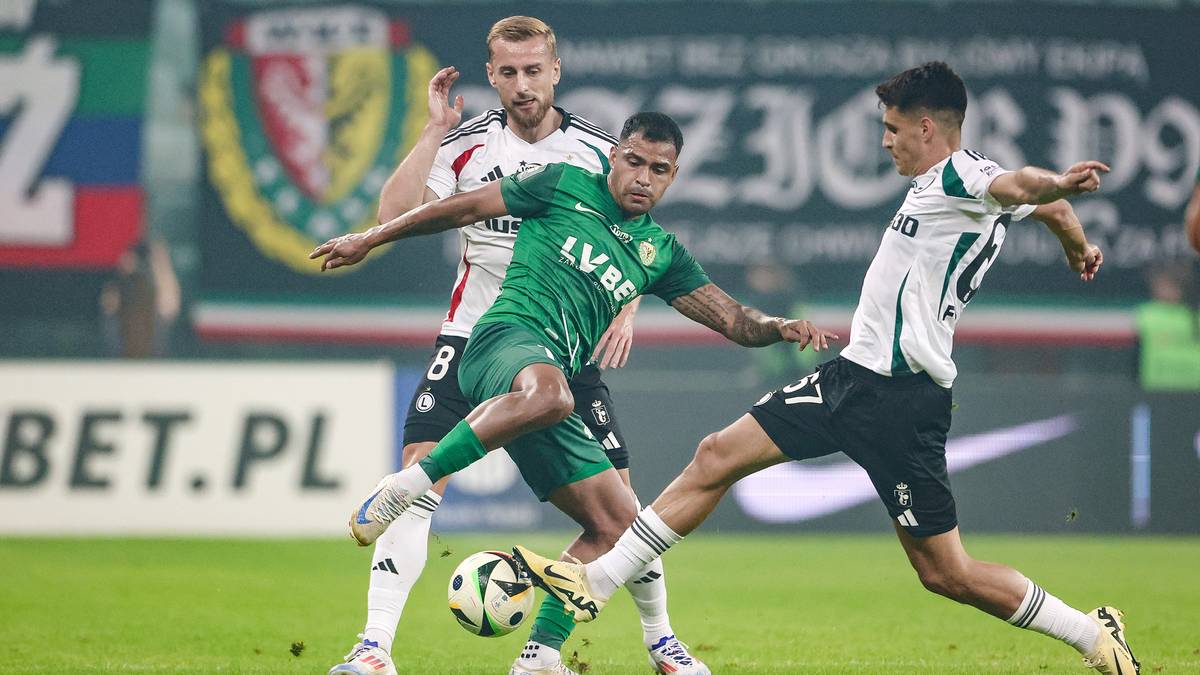 Piłkarze w akcji podczas meczu piłki nożnej, jeden z nich w zielonej koszulce pr&oacute;buje odebrać piłkę dw&oacute;m przeciwnikom w białych strojach.