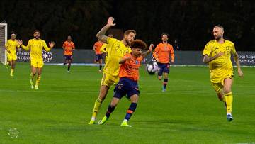 Dw&oacute;ch piłkarzy w ż&oacute;łtych strojach rywalizuje o piłkę na boisku piłkarskim, z innymi graczami w tle.