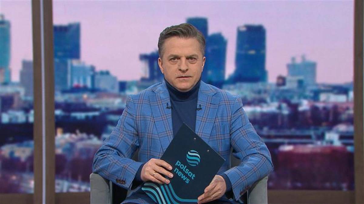 Bogdan Rymanowski siedzi w fotelu, trzymając w dłoniach tablet z logo Polsat News. W tle widoczna jest panorama miasta.