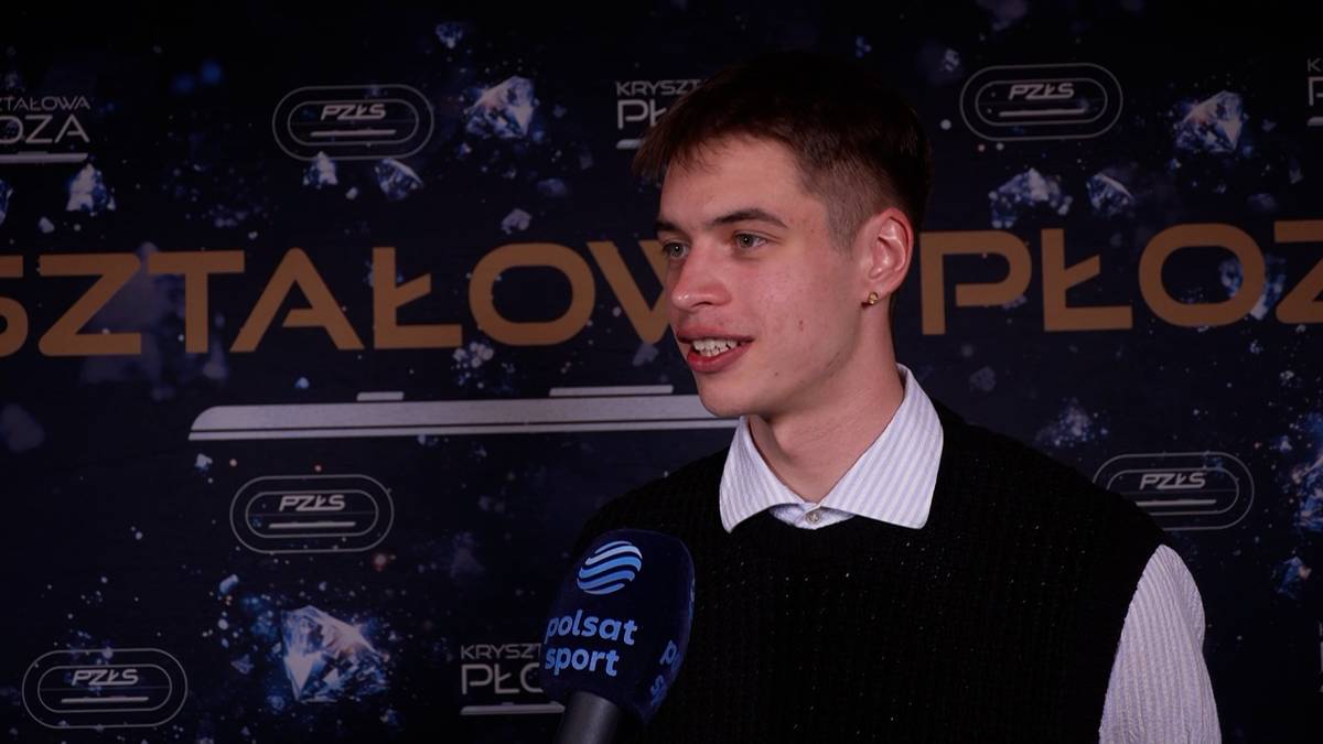 Młody mężczyzna w czarnej kamizelce i białej koszuli z kołnierzykiem m&oacute;wi do mikrofonu z logo Polsat Sport.